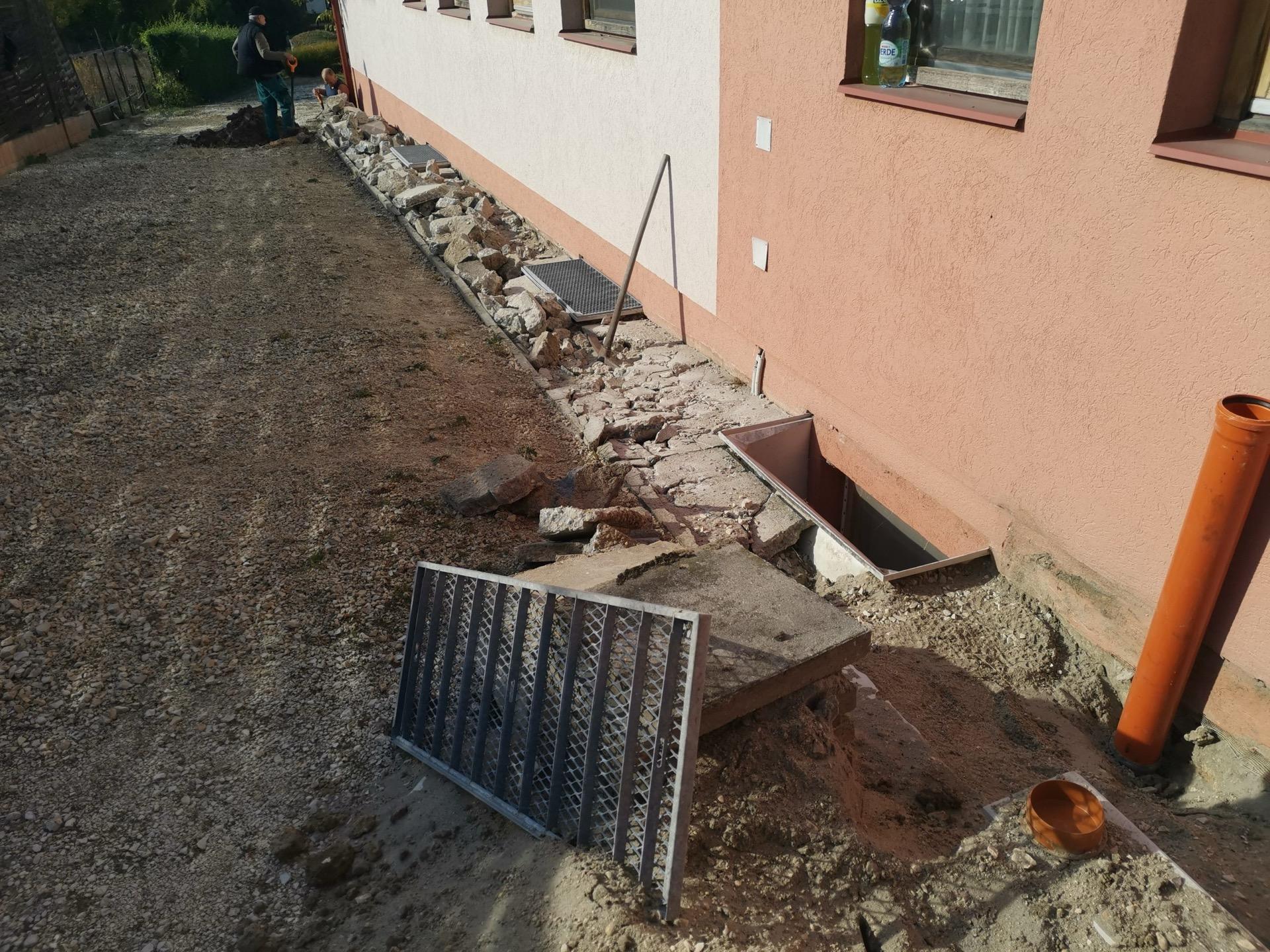 Munkaárok családi ház fala mellett: felbontott betonjárda, narancssárga dréncső és fémrácsok, a háttérben két dolgozó ás.