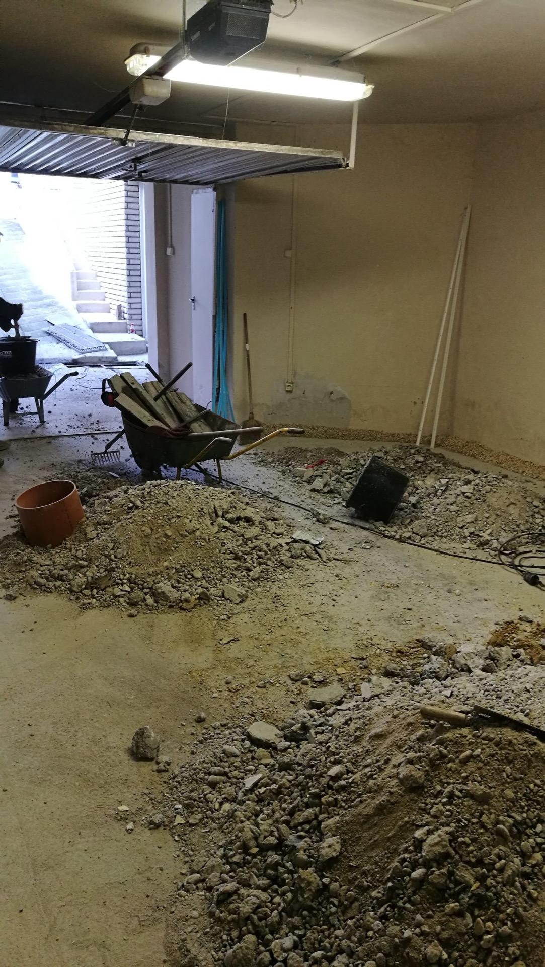 Felbontott aljzatú garázs nyitott szekcionált kapuval, kitermelt föld- és sittkupacokkal, talicskával.