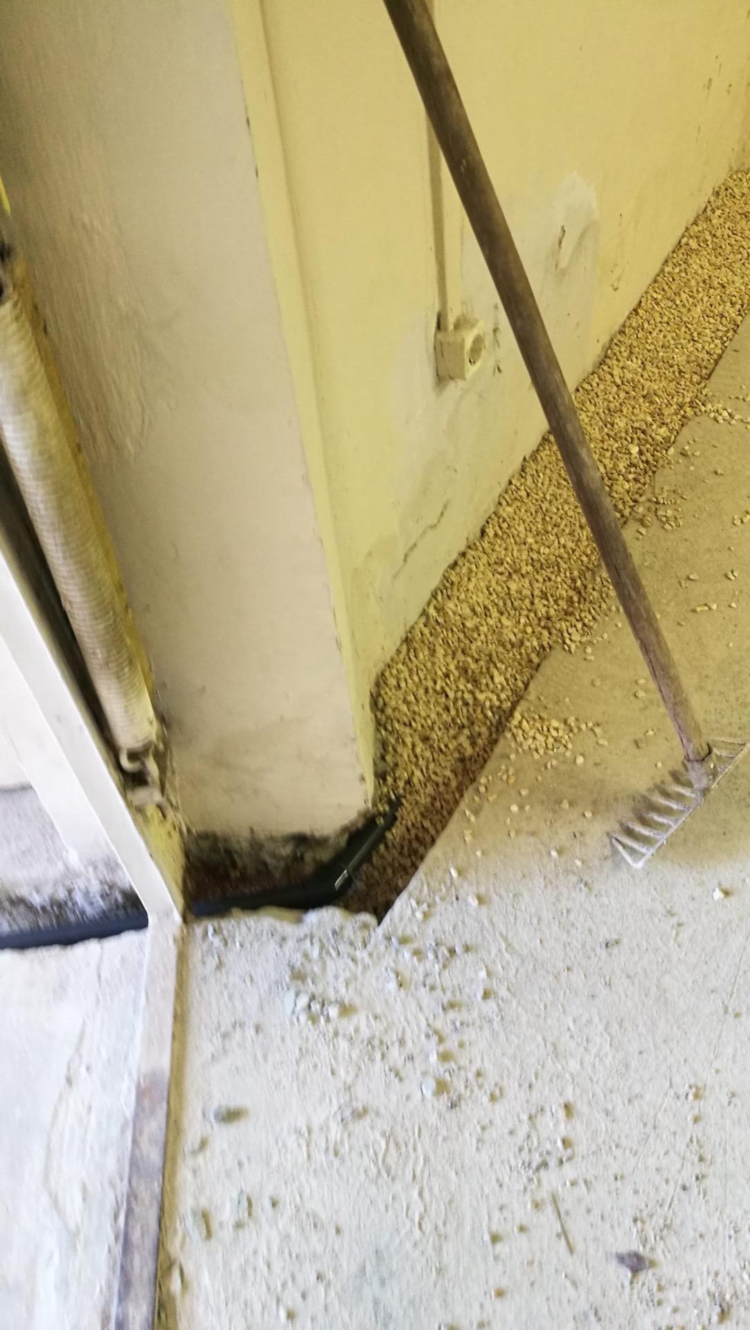 Fal tövébe vésett vezetékárok fekete csővel egy helyiség sarkában, a padlón szétterített kavics és gereblye.