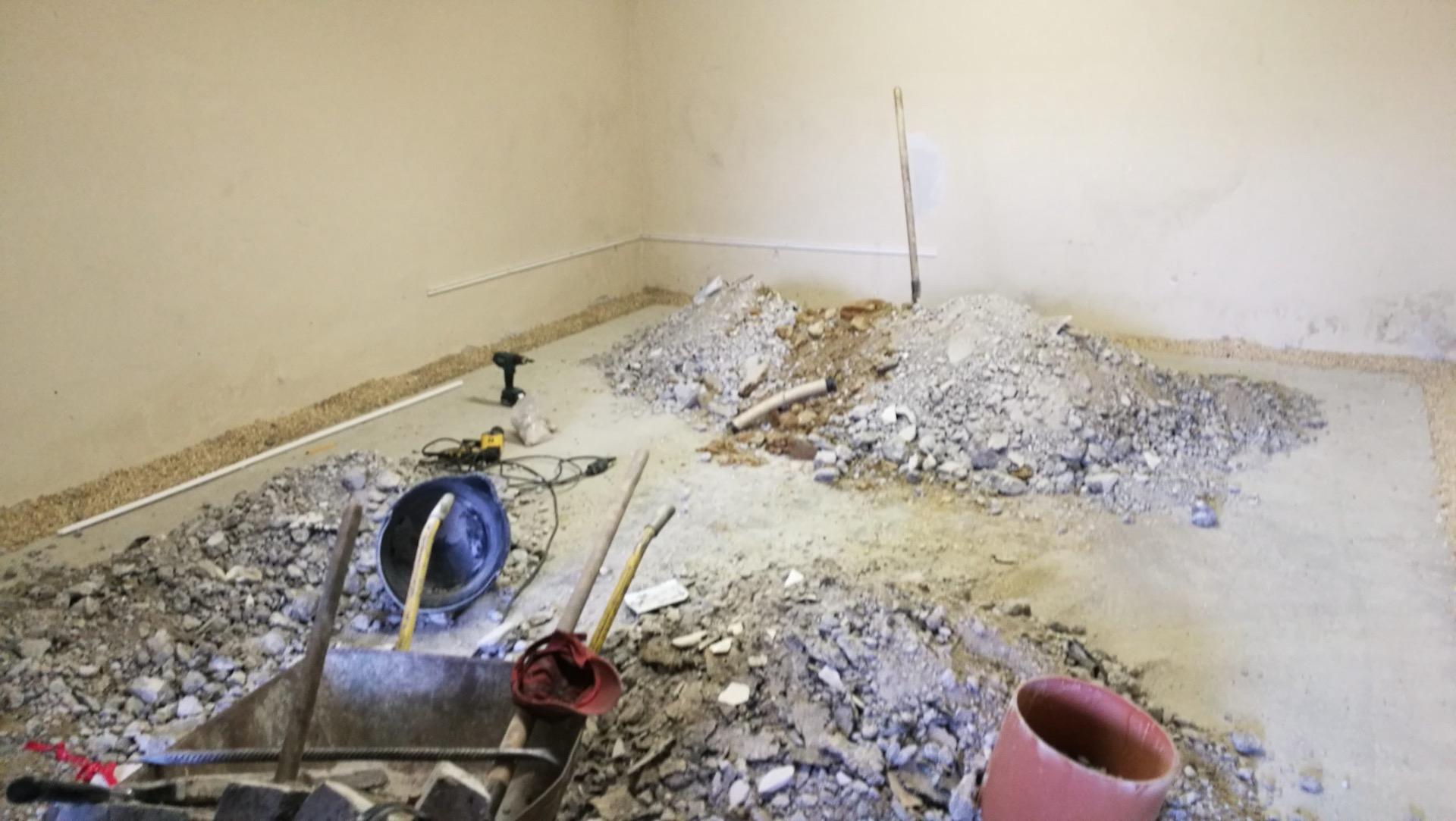 Felbontott padlójú helyiség: betontörmelék-kupac, a falak tövében kavicságy, előtérben talicska, védősisak és szerszámok.