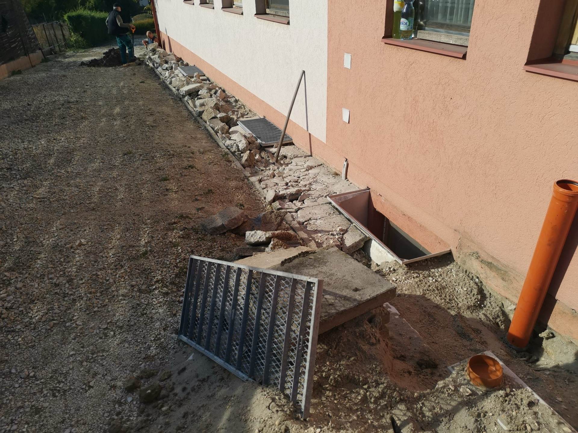 Munkaárok családi ház fala mellett: felbontott betonjárda, narancssárga dréncső és fémrácsok, a háttérben két dolgozó ás.